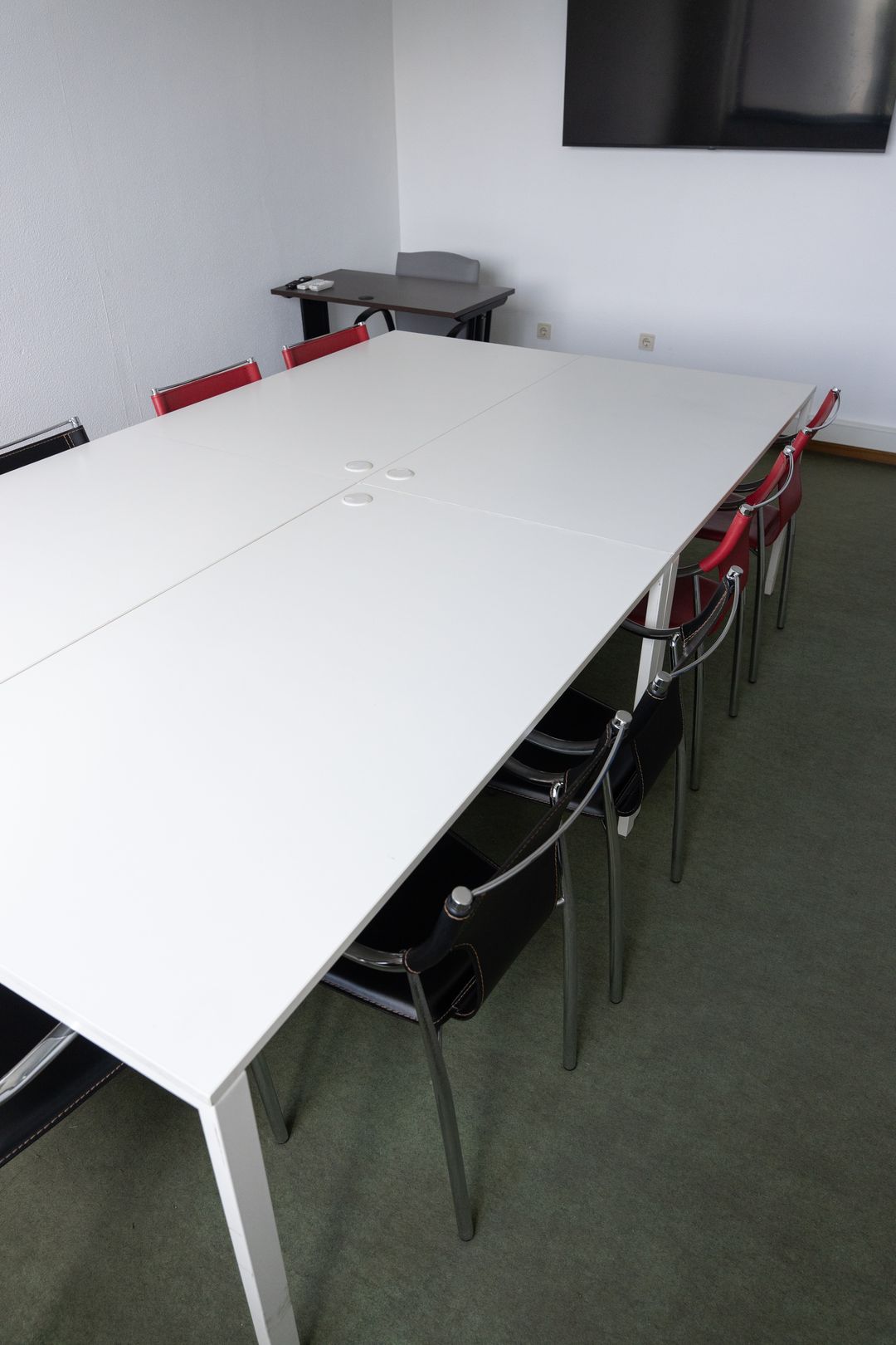 Mesa comprida com cadeiras vermelhas e pretas e a televisão ao fundo.
